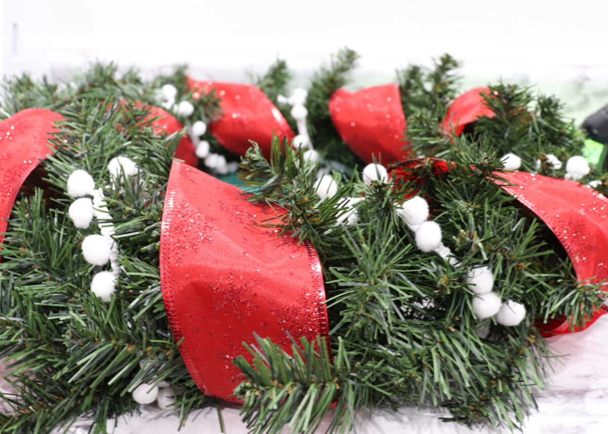 Image shows a faux pine wreath alternately wrapped with wide red ribbon and white pom-pom garland.
