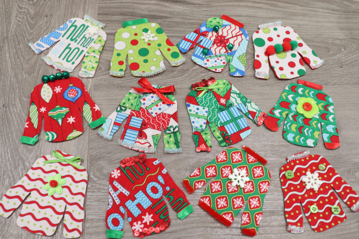 Image contains 12 small fabric “ugly sweaters” made from brightly patterned holiday fabric and decorated with embellishments like ribbon and buttons.