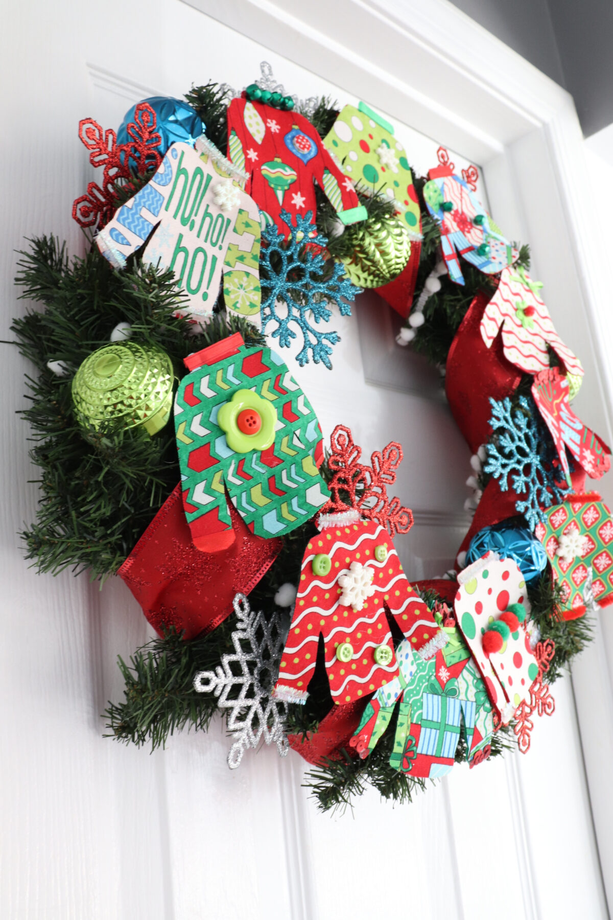 Image contains a colorful wreath made of small fabric “ugly sweaters” along with assorted ornaments an ribbon, hanging on a white door.
