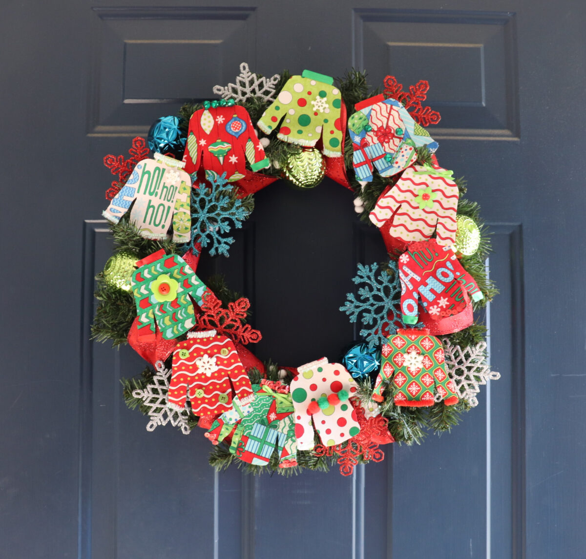 Image contains a colorful wreath made of small fabric “ugly sweaters” along with assorted ornaments an ribbon, hanging on a dark blue door.