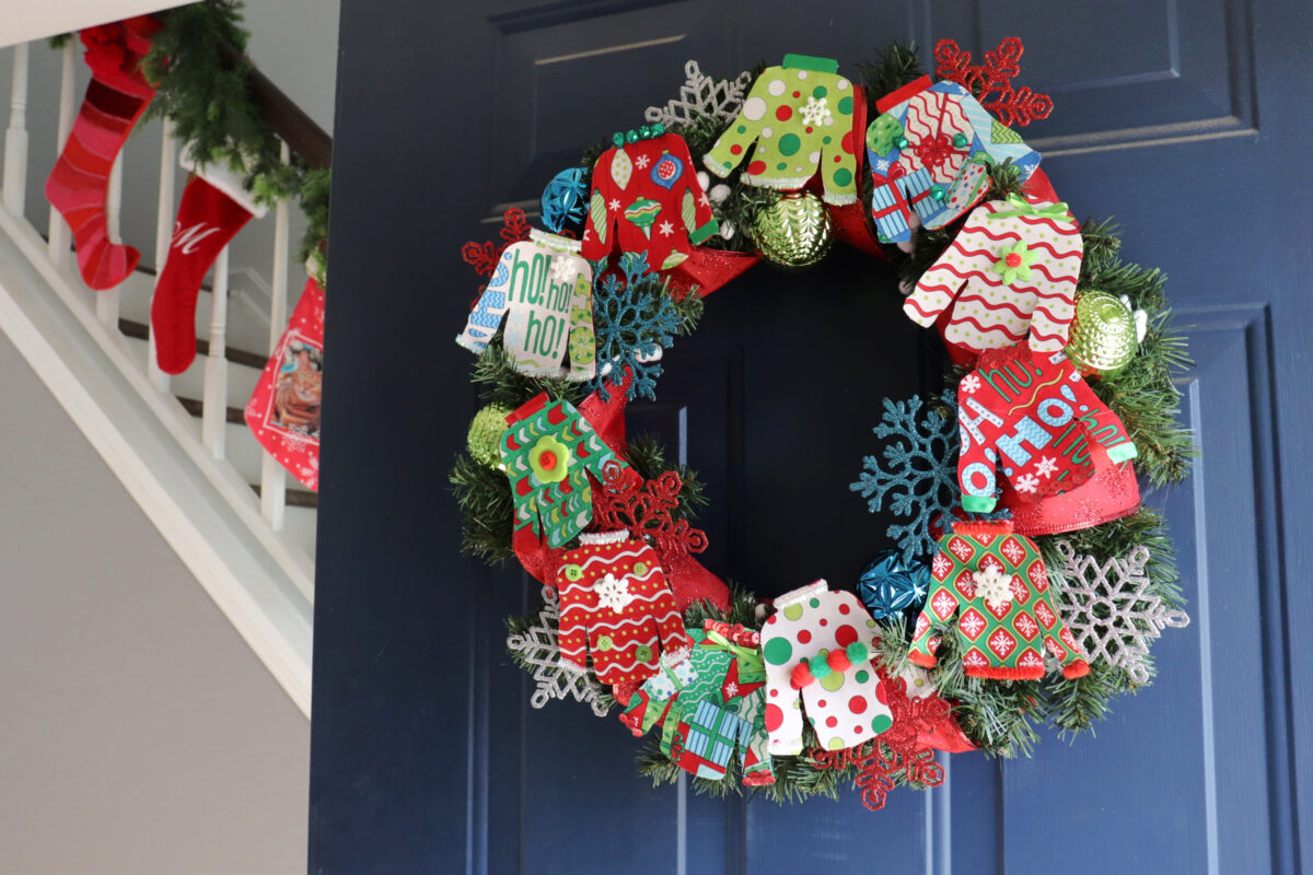 Image contains a colorful wreath made of small fabric “ugly sweaters” along with assorted ornaments an ribbon, hanging on a dark blue door. A staircase with several stockings extends out to the left past the doorl.