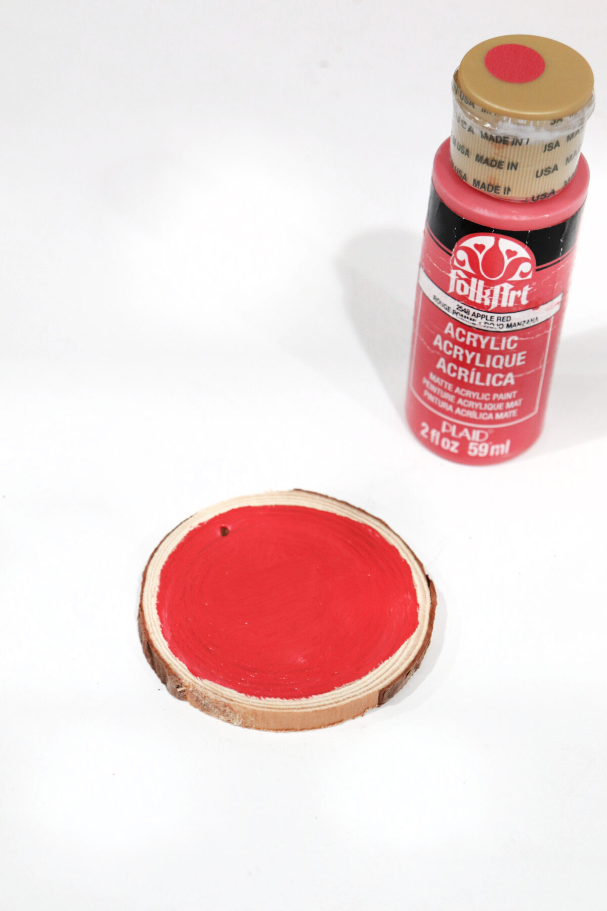 Image contains a wood slice painted red, sitting on a white tabletop with a bottle of FolkArt red acrylic paint nearby.
