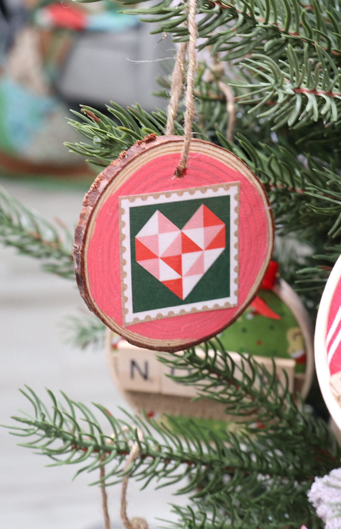 Image contains a wood slice ornament painted salmon pink with a quilted heart design decoupaged on top. It hangs on a fake tree with a piece of twine.