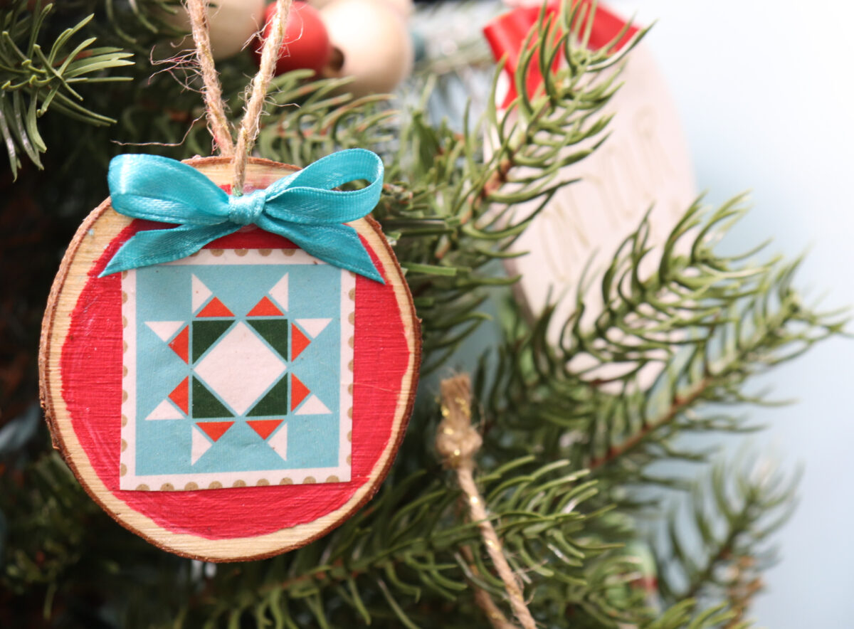 Image contains an ornament made from a wood slice that has been painted red with a quilt star image decoupaged on top. It hangs on a faux tree from a twine hanger with a teal bow.