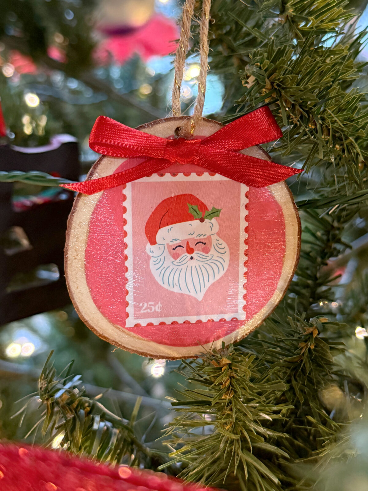 Image contains a wood slice ornament painted pink with a cartoon image of Santa on top. It hangs from a piece of twine on an artificial tree.