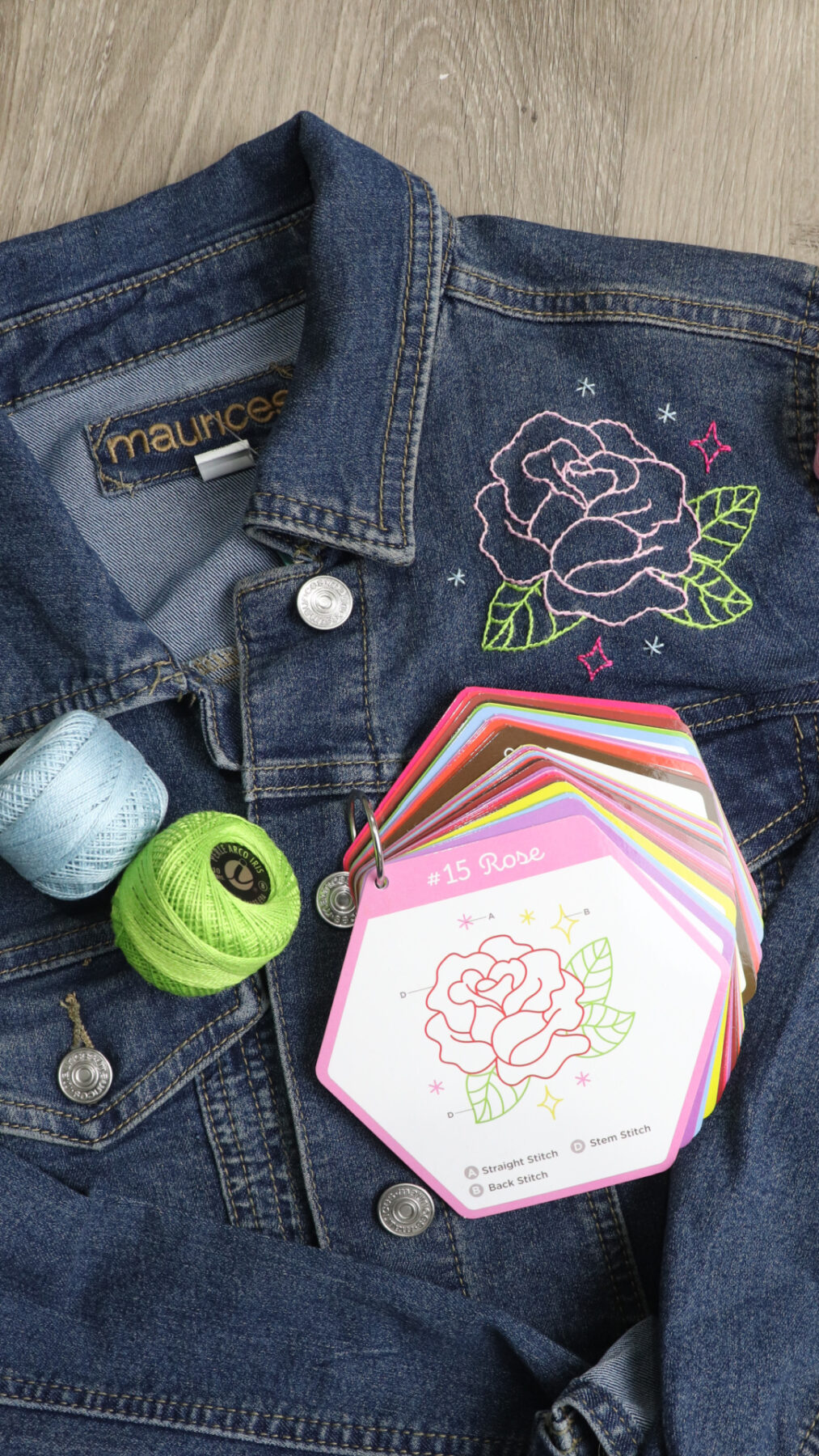 Image contains a denim jacket with an embroidered rose. Two spools of floss and a ring of stitch guide cards sit on top.