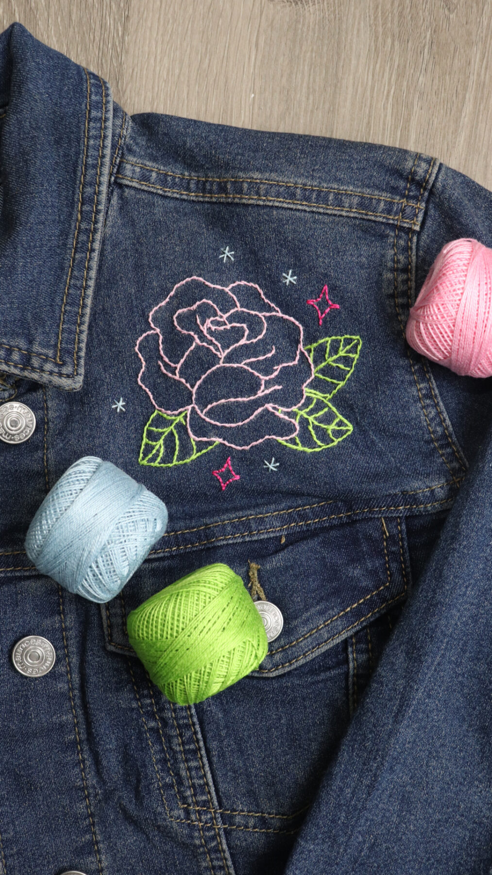 Image contains the front shoulder area of a denim jacket that has been embellished with an embroidered rose. Spools of blue, pink, and green perle cotton lay on top.
