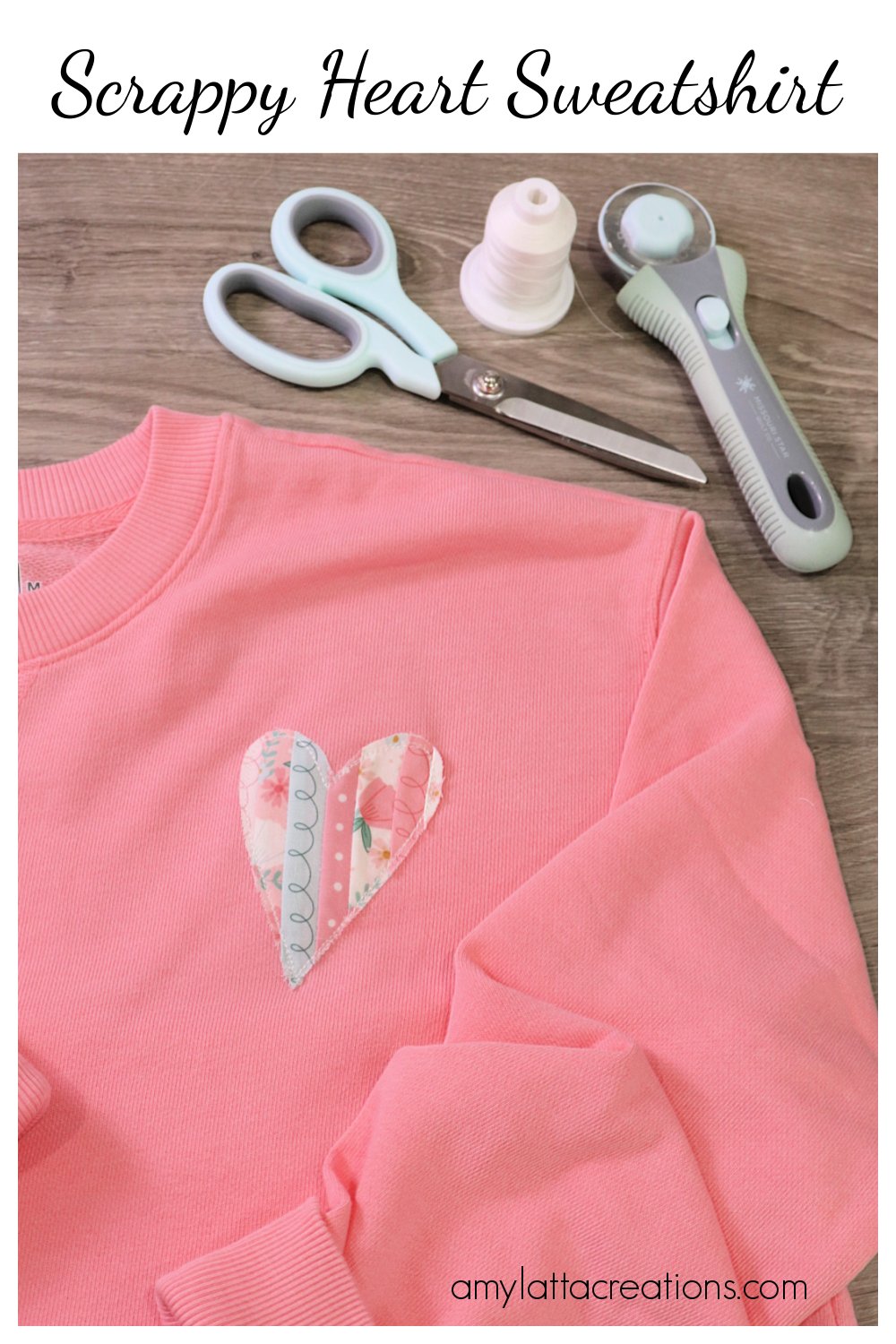 Image contains a coral colored sweatshirt embellished with an applique fabric heart. It says on a wooden table near a pair of scissors, a spool of thread, and a rotary cutter.