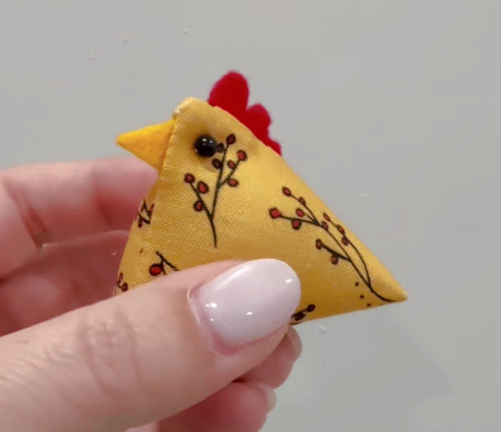 Image contains Amy’s hand holding the finished tiny chicken pin cushion, complete with small beads for eyes.