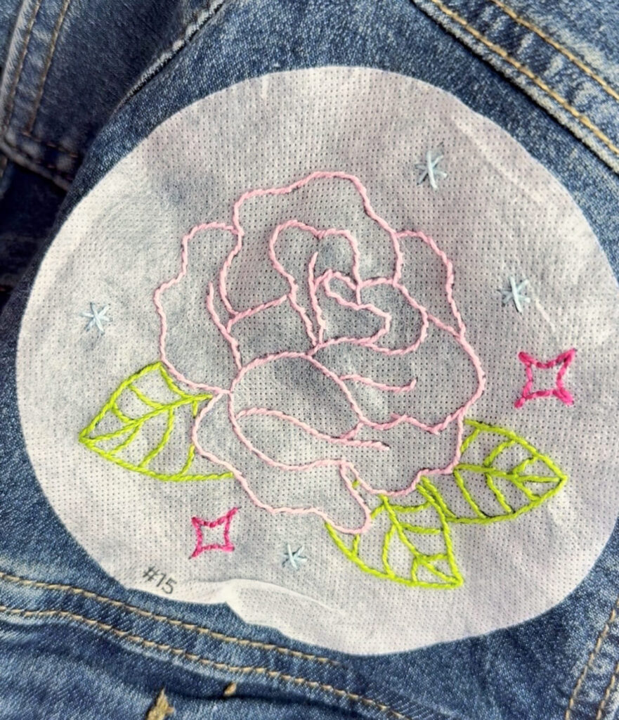 Image contains a close up view of the embroidered rose design, stitched on top of the round embroidery paper with pink, blue, and green floss.