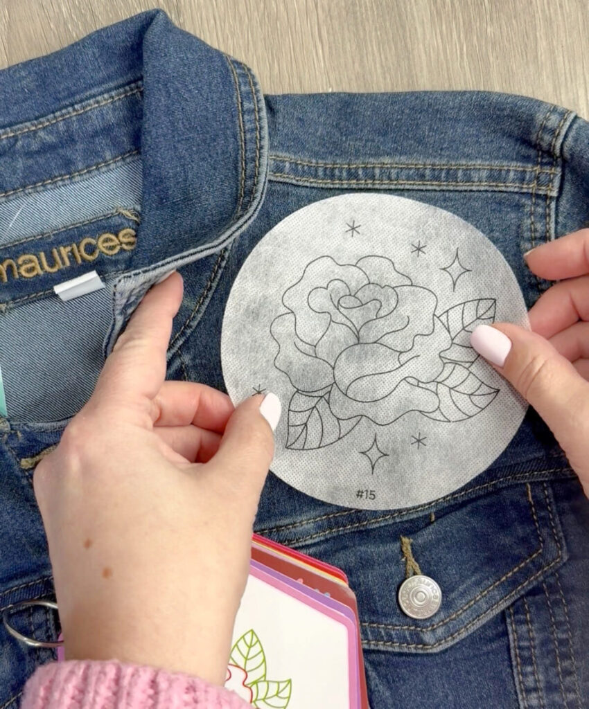 Image contains Amy’s hands affixing a circle with a printed design to the front of a denim jacket.