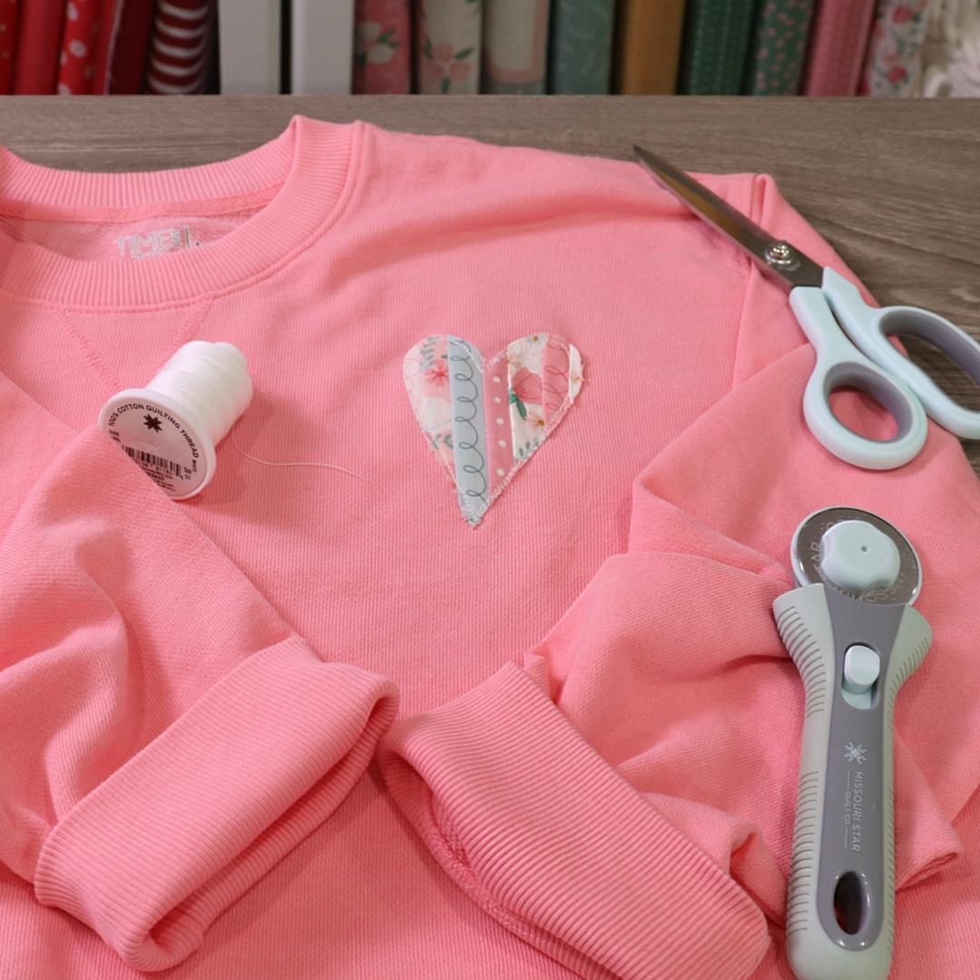 Image contains a wooden table holding a pink sweatshirt embellished with a striped fabric heart. Scissors, a rotary cutter, and white thread lay nearby.