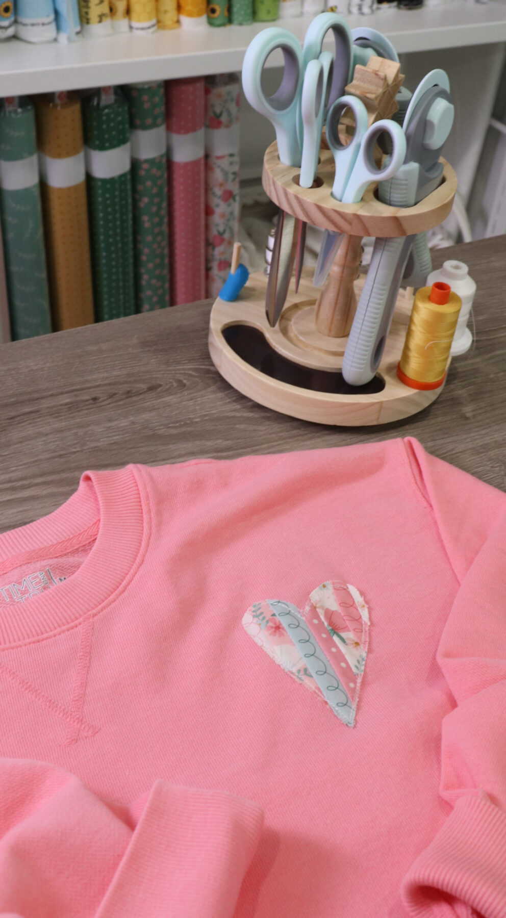 Image contains a pink sweatshirt with an applique fabric heart laying on a wooden table. A wooden tool caddy sits nearby with scissors, thread, and rotary cutters.