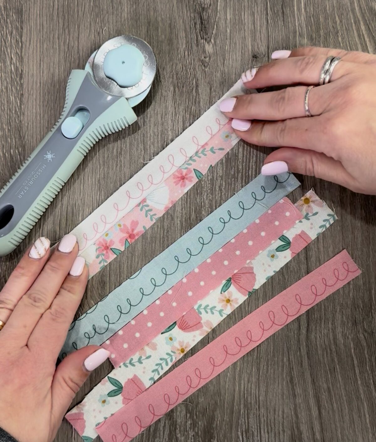 Image contains six strips of fabric laying on a wooden desk along with a rotary cutter. Amy’s hands hold two of the strips, which have been sewn together.