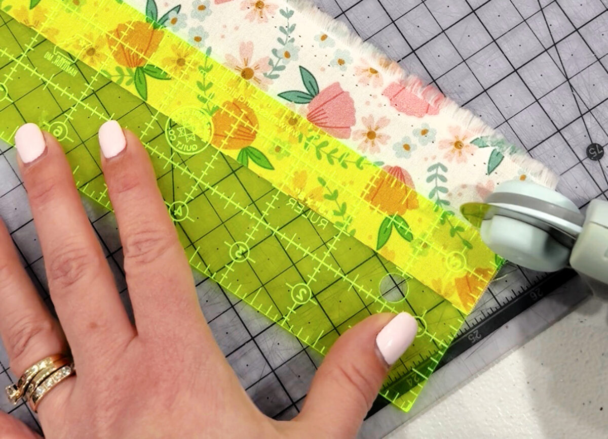Image contains a piece of white and pink floral fabric laying on a clear cutting mat. Amy’s hand holds a yellow ruler on top of the fabric, and a rotary cutter is positioned at the bottom.