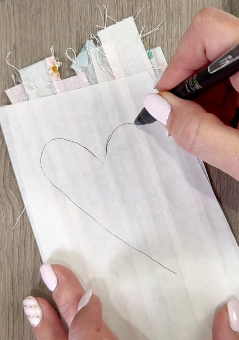 Image contains Amy’s hand holding a black pen and drawing a heart shape onto the back of a fabric rectangle with fusible adhesive on top.