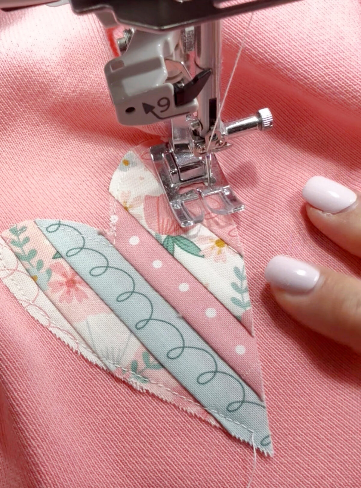 Image shows a closeup view of a pink sweatshirt on a sewing machine. The needle is sewing an applique heart in place.