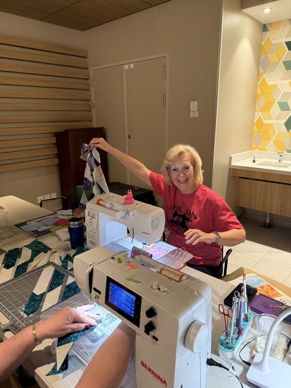 Image contains a smiling woman sitting at a sewing machine and holding up a partly finished quilt top.
