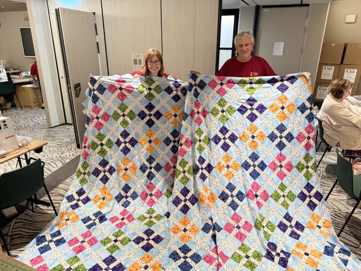 Image contains a woman and a man, each holding an identical finished quilt top.