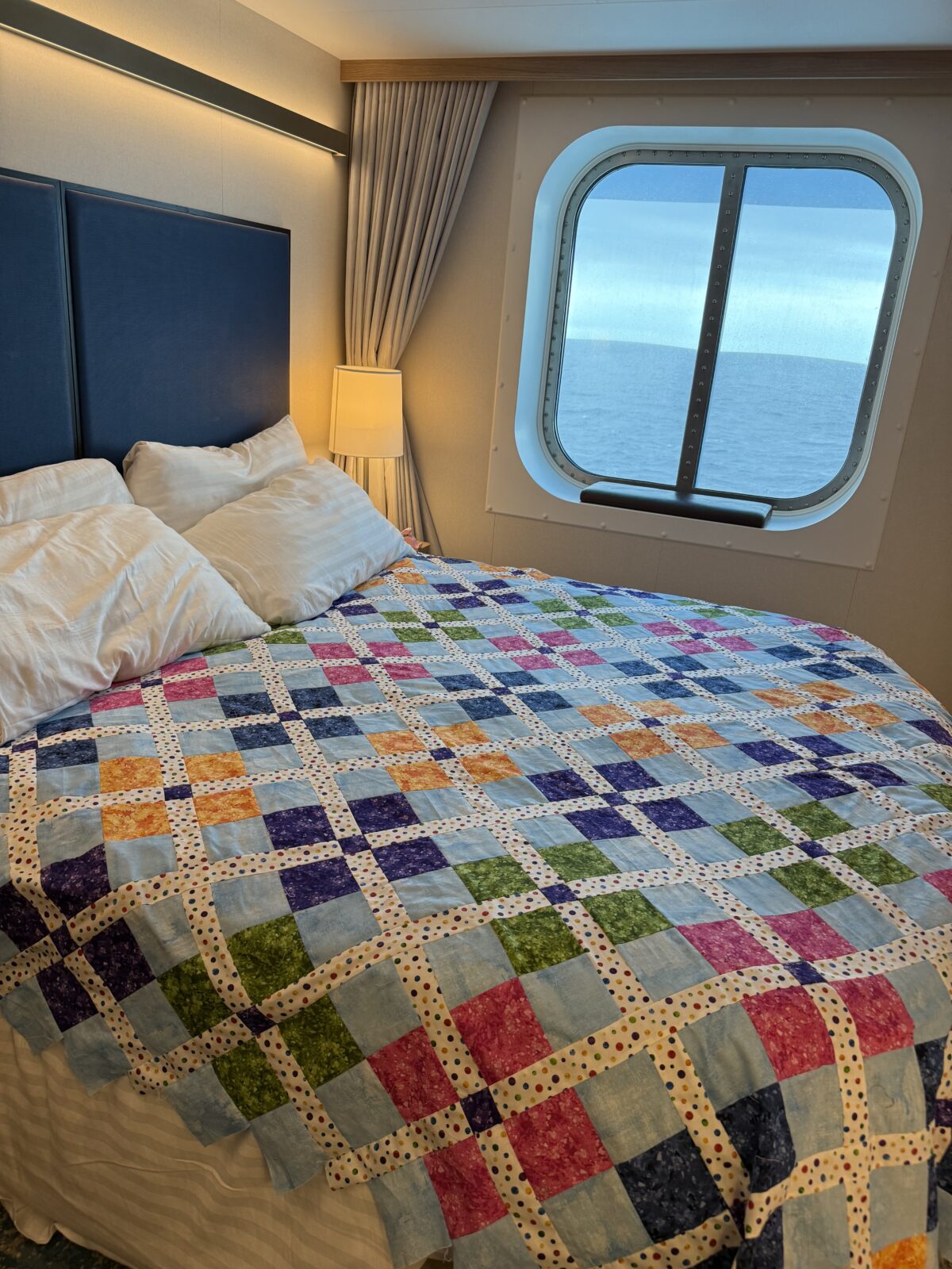 Image contains a cruise ship state room with a window. The bed is covered with a colorful quilt top.