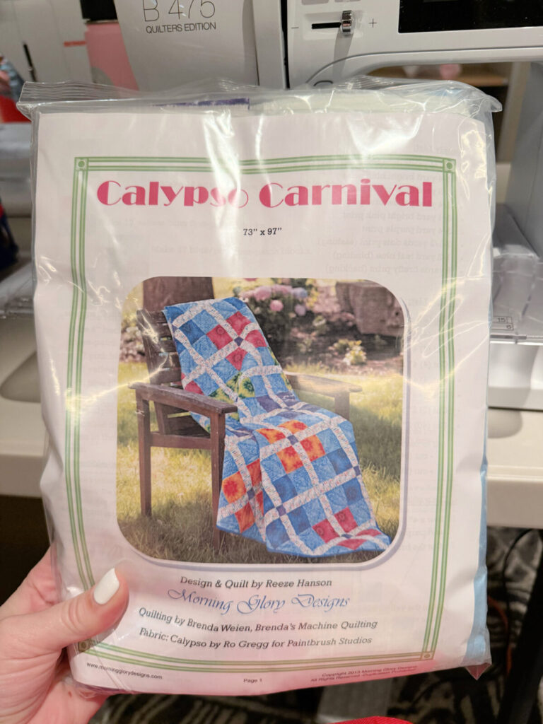 Image contains Amy’s hand holding the Calypso Carnival Quilt Kit. A Bernina sewing machine is in the background.