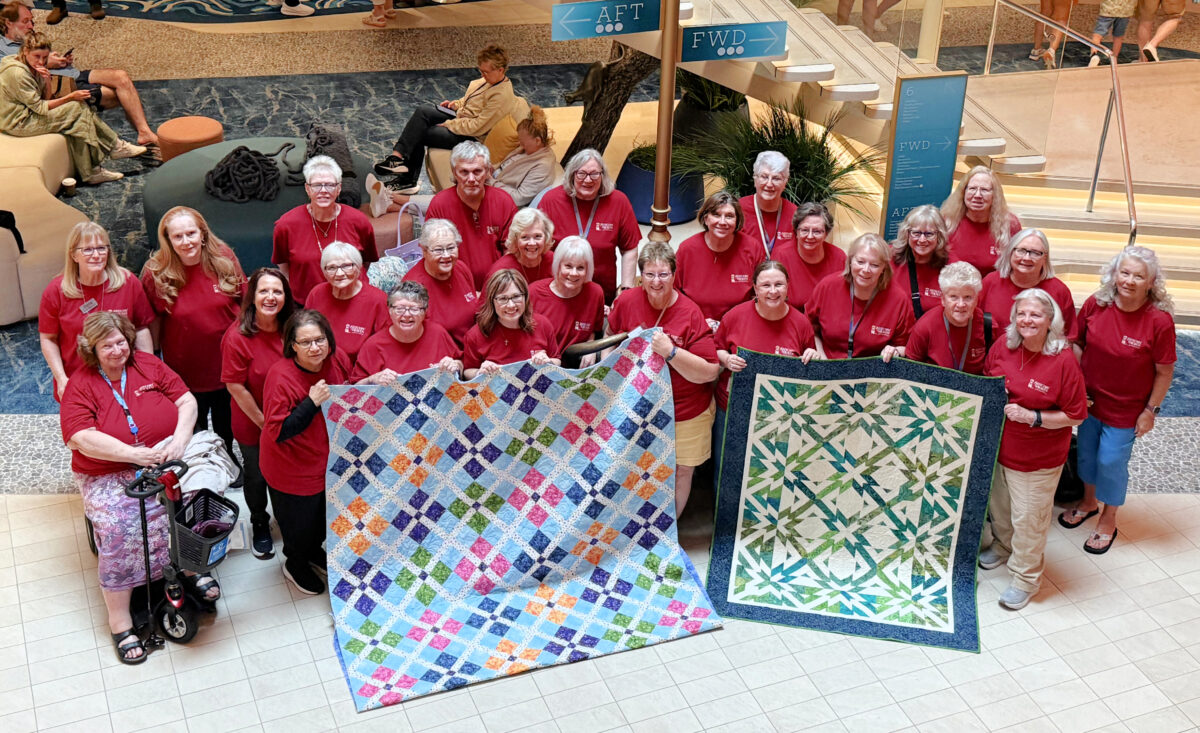 Image contains a group of quilters in red shirts holding up two finished quilts, standing on the 5th deck of the Icon of the Seas.