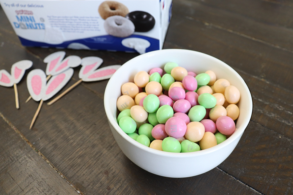 Image contains a bowl of pastel m&m candies sitting on a dark wood table. Bunny ear picks and a pack of mini powdered donuts sit nearby.