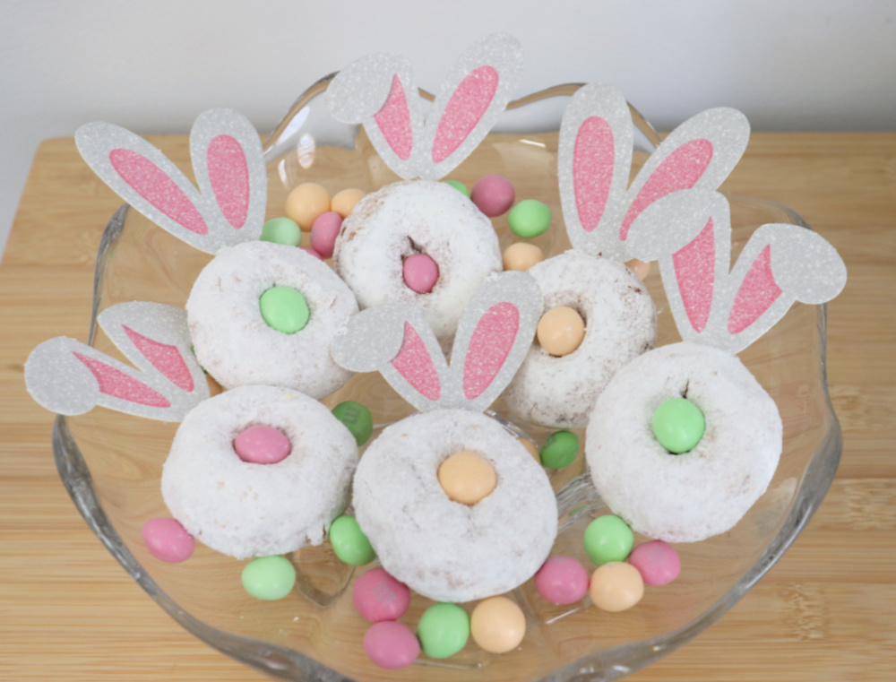 Image contains six mini bunny donuts arranged in a glass serving dish with pastel candies scattered around.