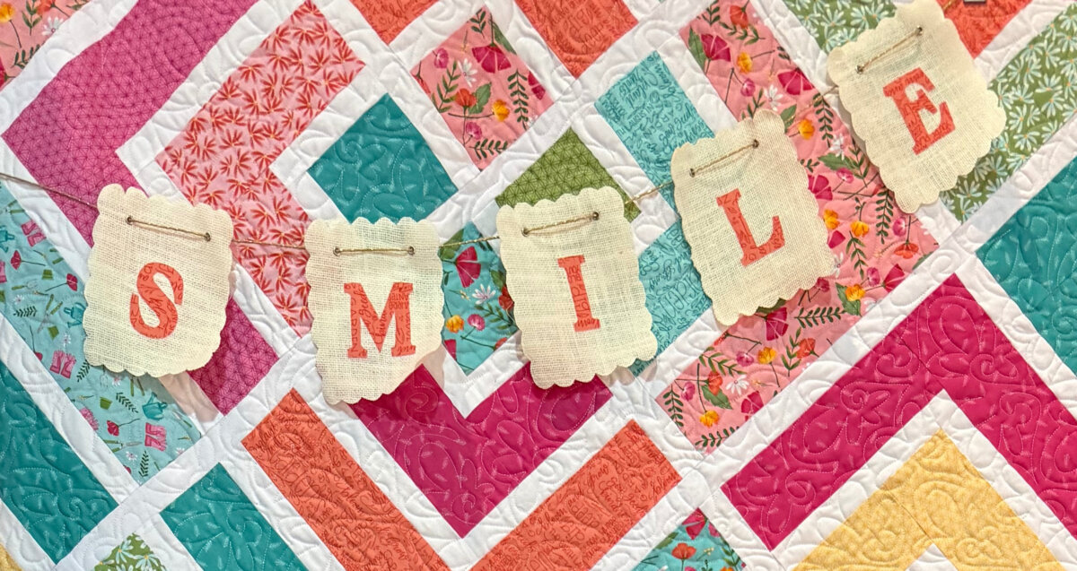Image contains a flag banner reading, “SMILE” in orange fabric, hanging in front of a bright quilt.