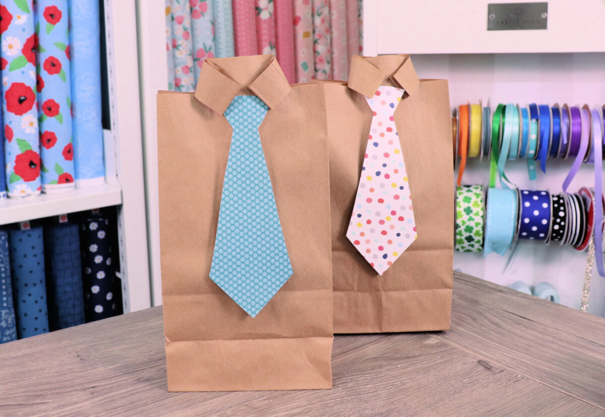 Image contains two completed gift bags on a wooden table, with ribbon and fabric in the background.