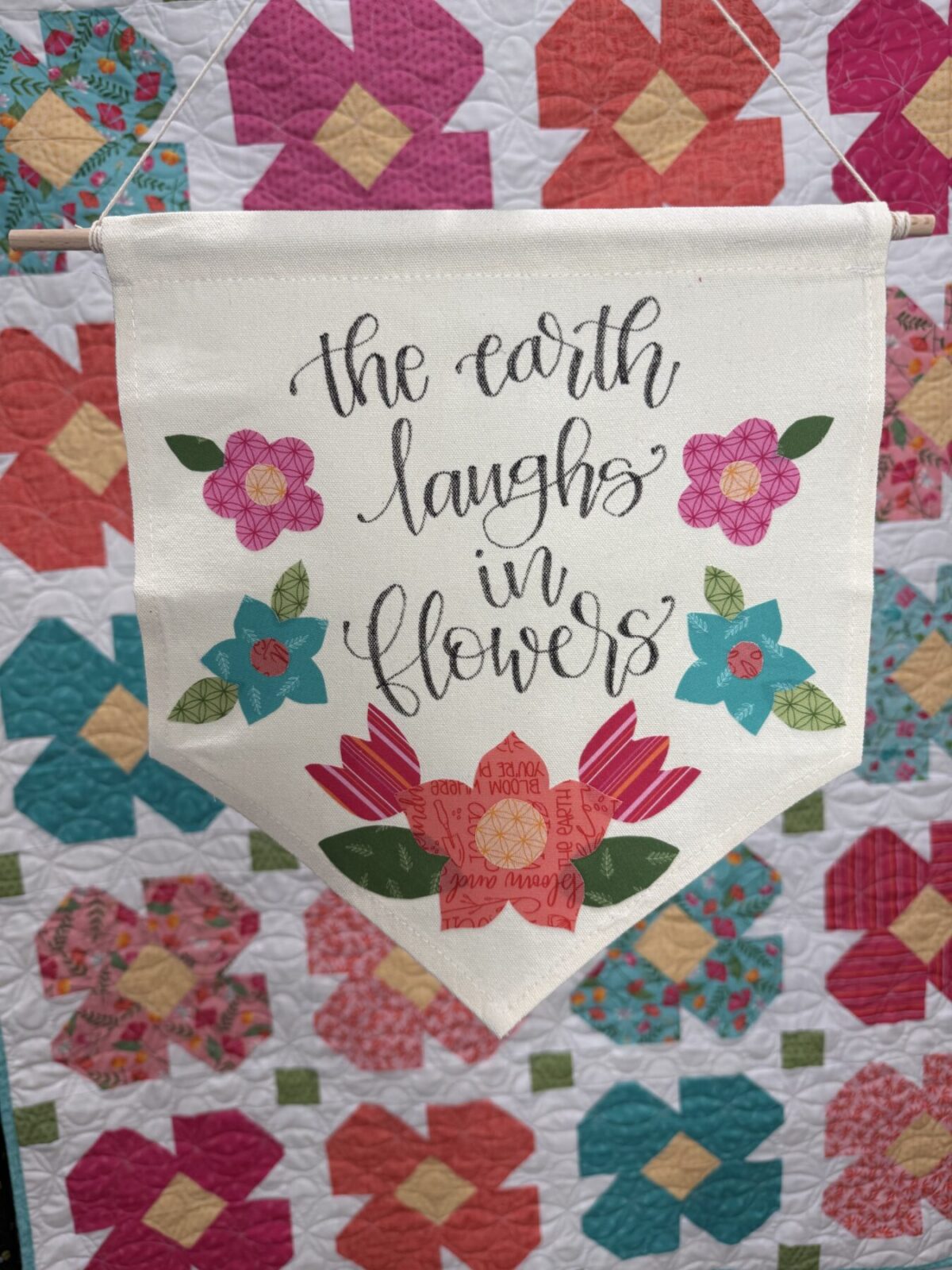 Image contains a canvas banner with applique fabric flowers and a hand lettered phrase in front of a floral quilt.
