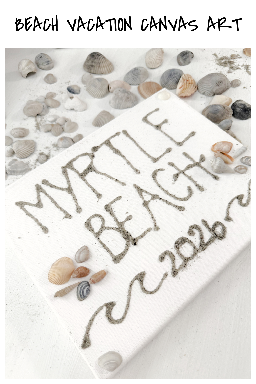 Image contains a white canvas with “Myrtle Beach 2026” written on it in sand. Several small shells decorate the canvas, which sits on a white table covered with more shells.