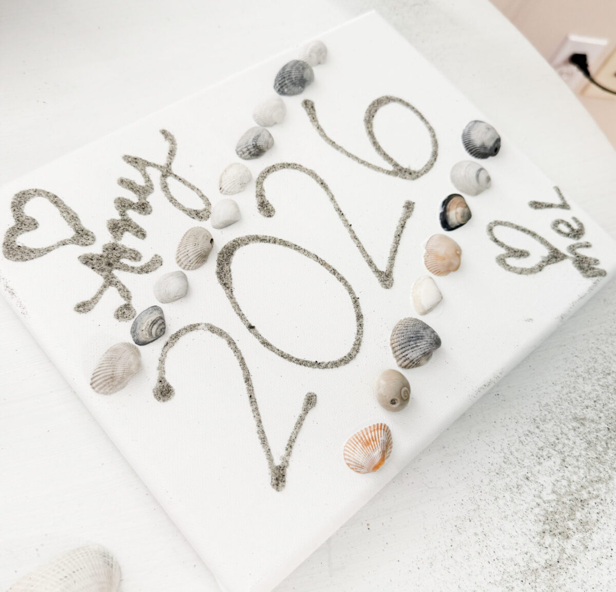 Image contains a white canvas with names, hearts, and “2026” written in sand. Two lines of shells are on either side of the date. The canvas sits on a white table.