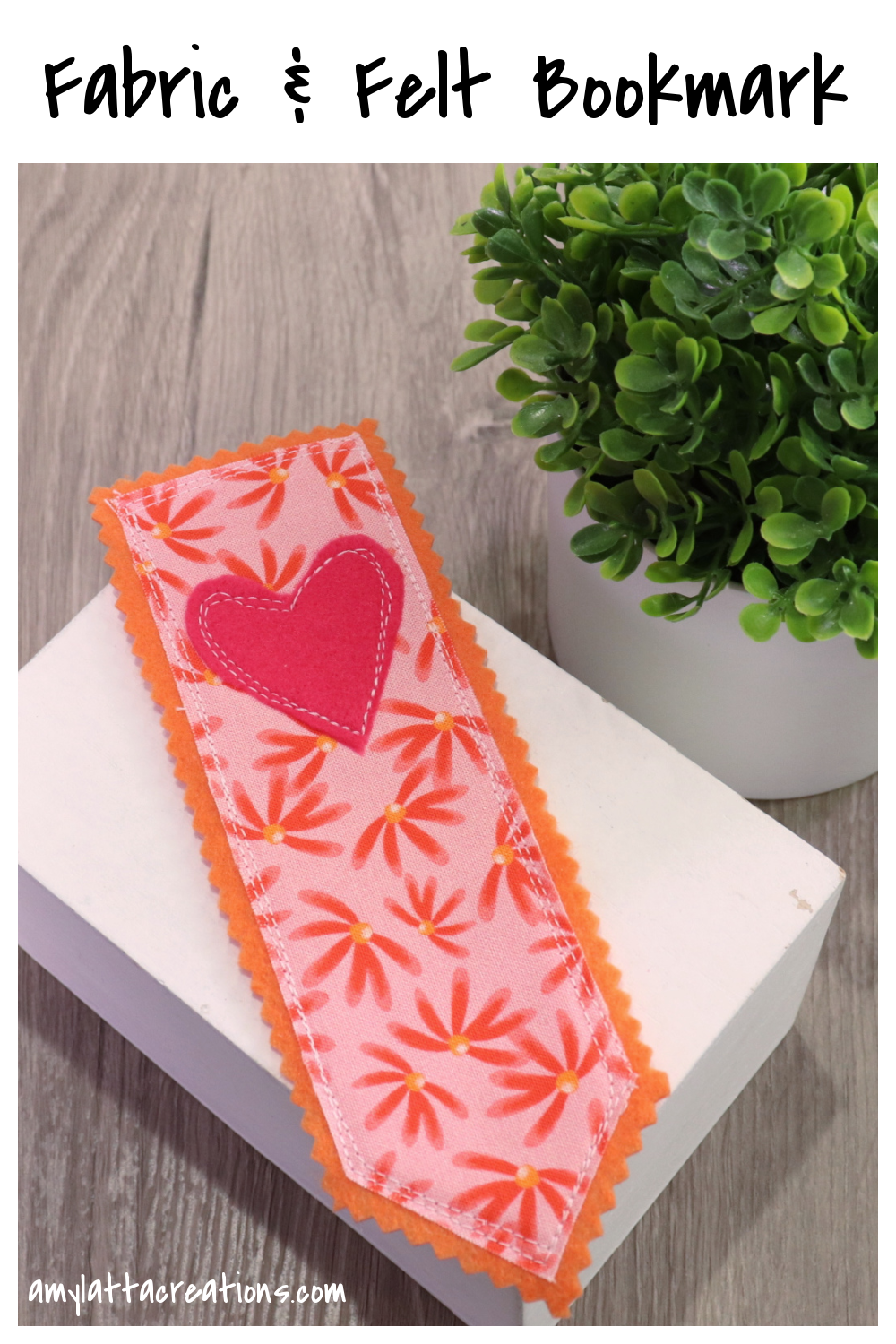 Image contains a bookmark made of orange felt and pink fabric with daisies. A pink felt heart is sewn on top. It sits on a small white box next to a faux plant on a wooden table.