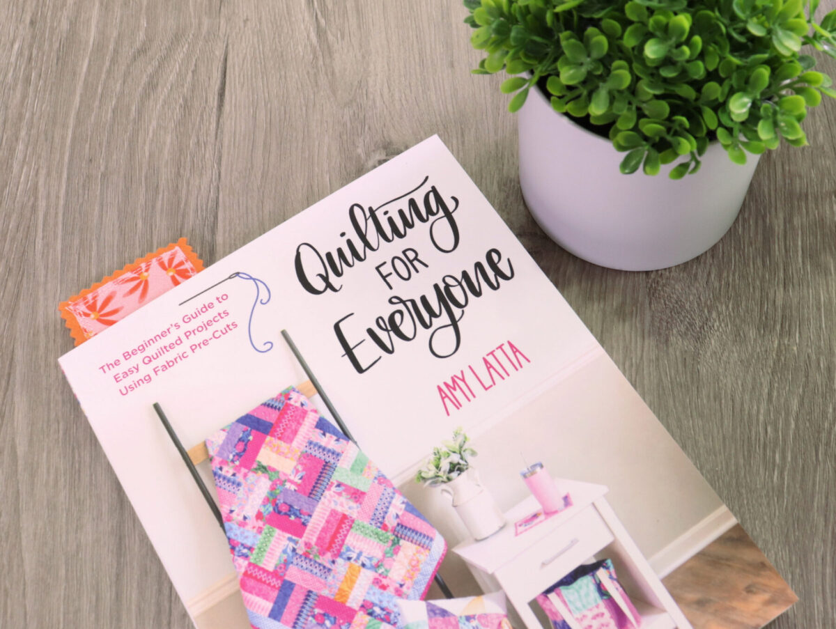 Image contains the book Quilting for Everyone by Amy Latta with a fabric bookmark sticking out from the top. It sits on a wooden desk next to a plant.