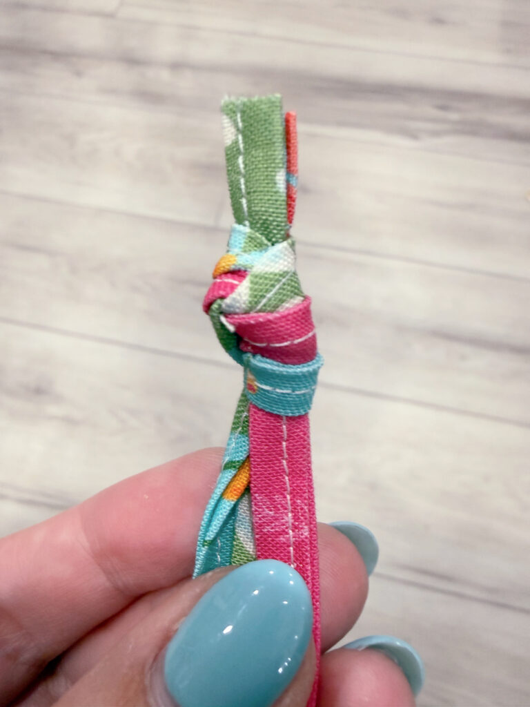 Image contains Amy’s hand holding three fabric strips knotted together at the top.