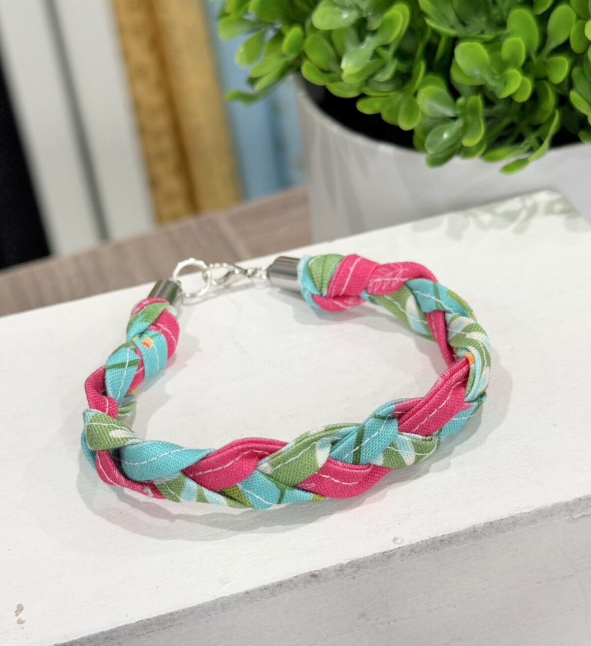 Image contains the completed bracelet made from braided fabric strips, sitting on a white platform next to a plant.