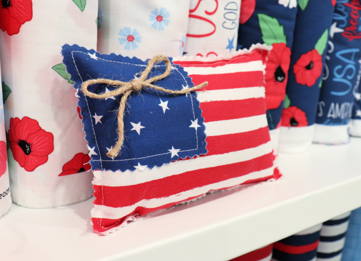 Image contains a small flag pillow made from patriotic fabrics, sitting on a shelf in front of fabric bolts.