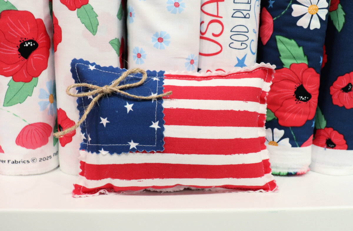 Image contains a small flag pillow made from patriotic fabrics. It sits on a shelf in front of other red, white, and blue fabric.