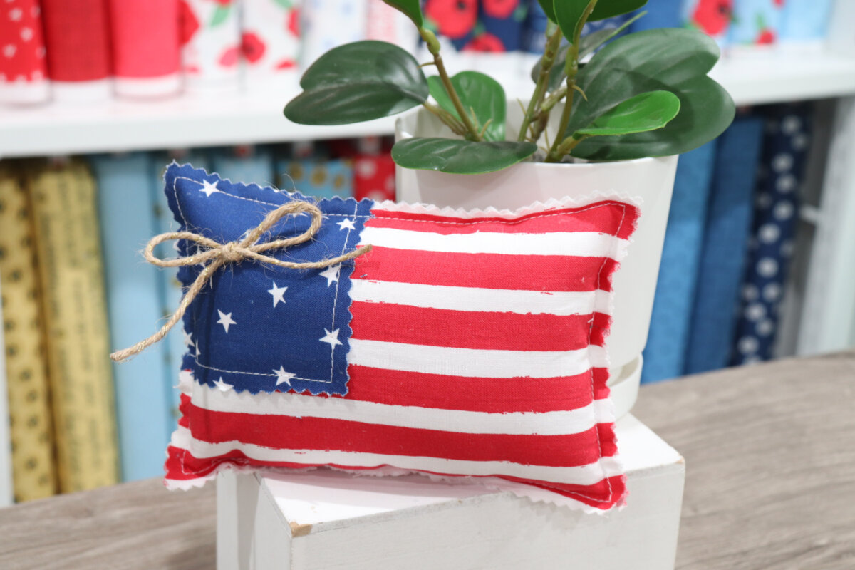 Image contains a small flag pillow made from red, white, and blue fabrics. It sits next to a faux plant on a small white box.