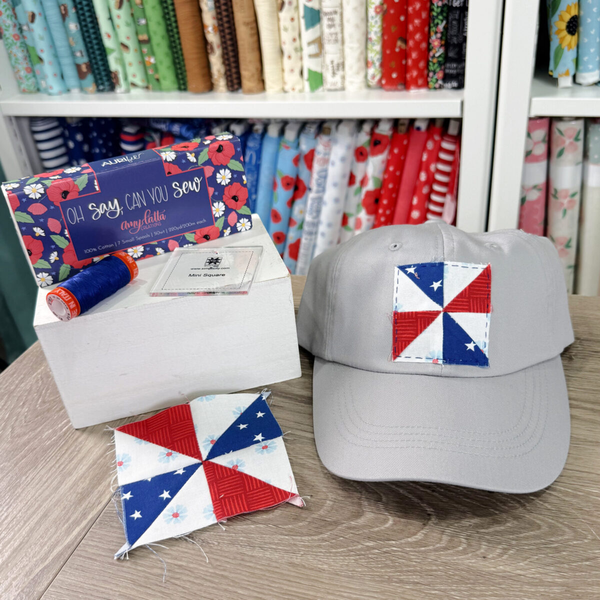 Image contains a grey hat decorated with a patriotic mini pinwheel quilt block. It sits next to a thread collection and another quilt block on a wooden desk.