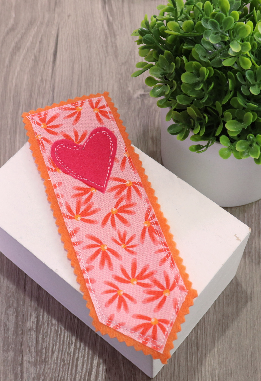 Image contains a pink and orange bookmark made of felt and fabric, decorated with daisies and a heart.