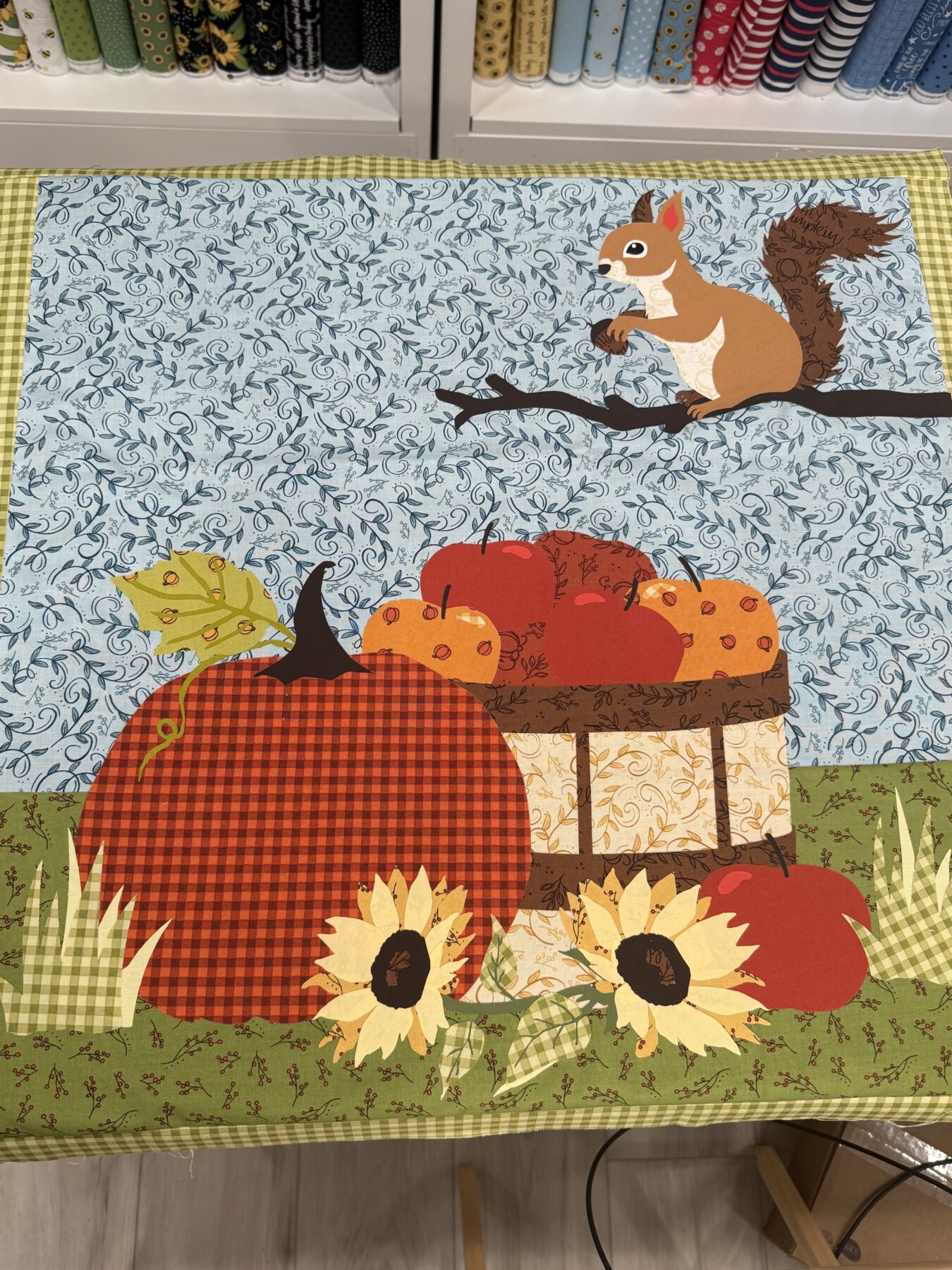 Image contains a quilt center with an applique fall scene featuring a squirrel holding a nut on a branch, a pumpkin, a barrel of pumpkins and apples, and sunflowers.