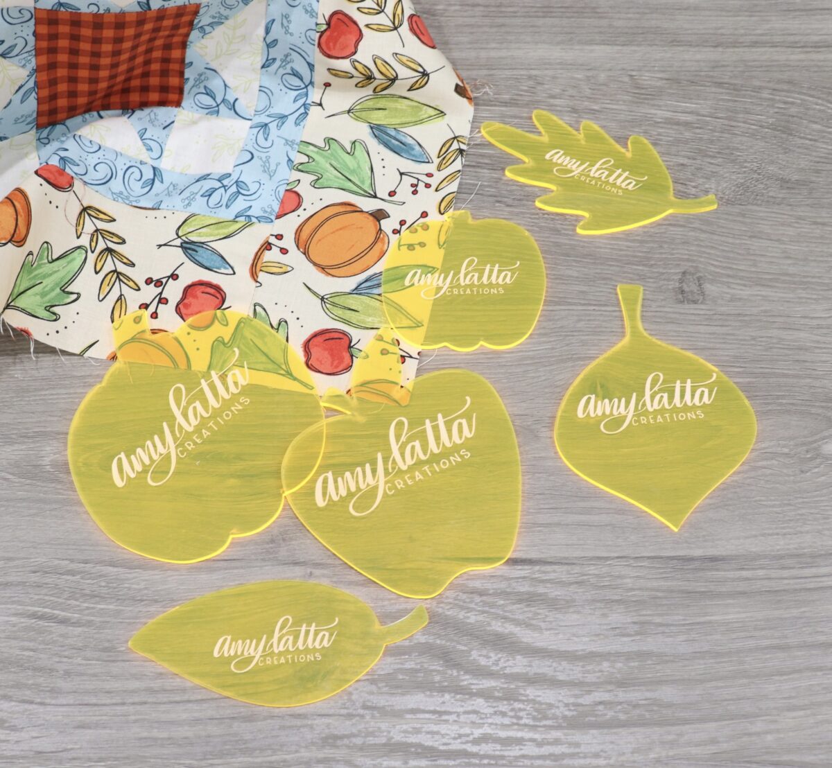 Image contains the six templates in the Harvest Home set: two pumpkins, three leaves, and an apple. They sit on a wooden desk with a quilt top made of Harvest Home fabric.