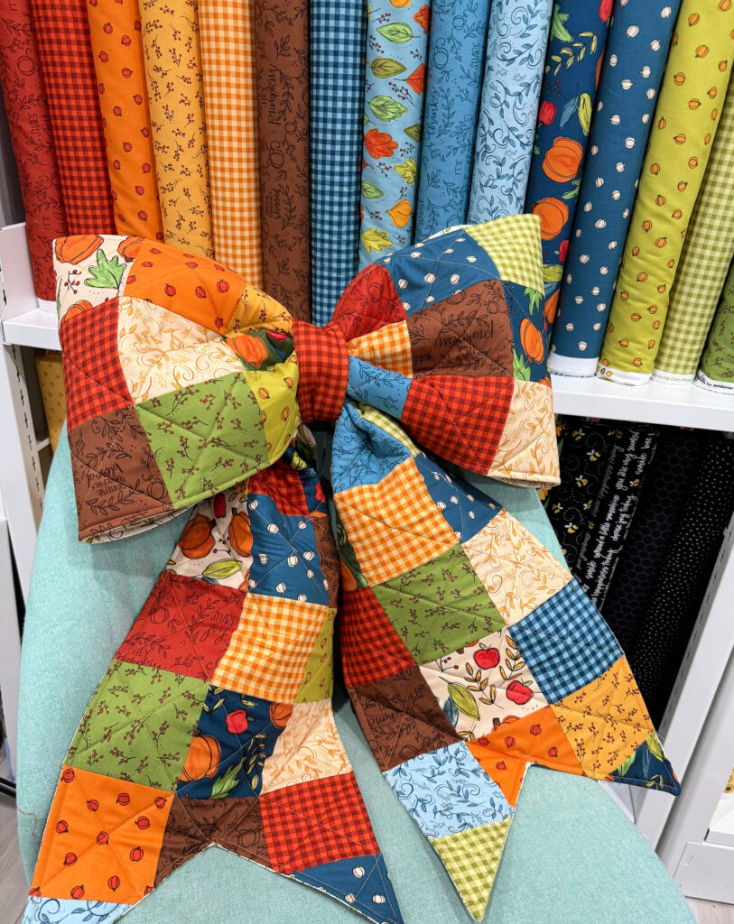 Image contains a large patchwork fabric bow sitting on a teal chair in front of bolts of fall fabric.