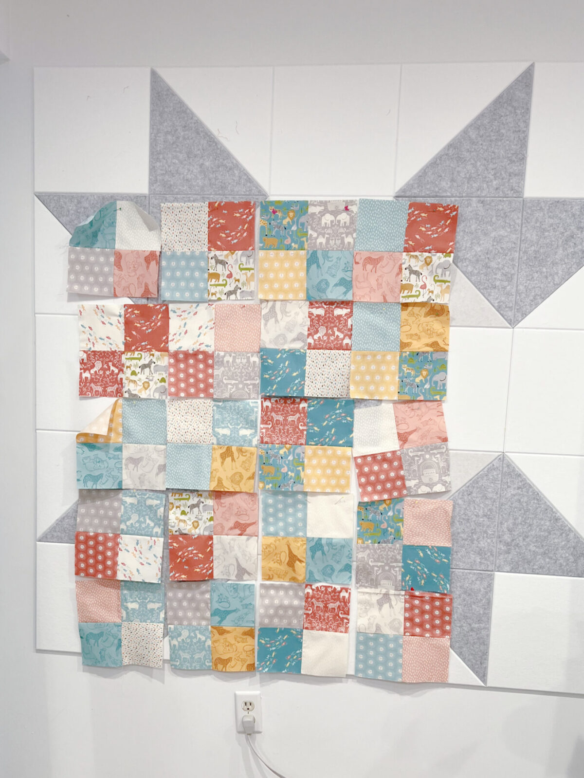 Image contains a white and grey felt quilt wall with multicolored quilt blocks pinned to it.
