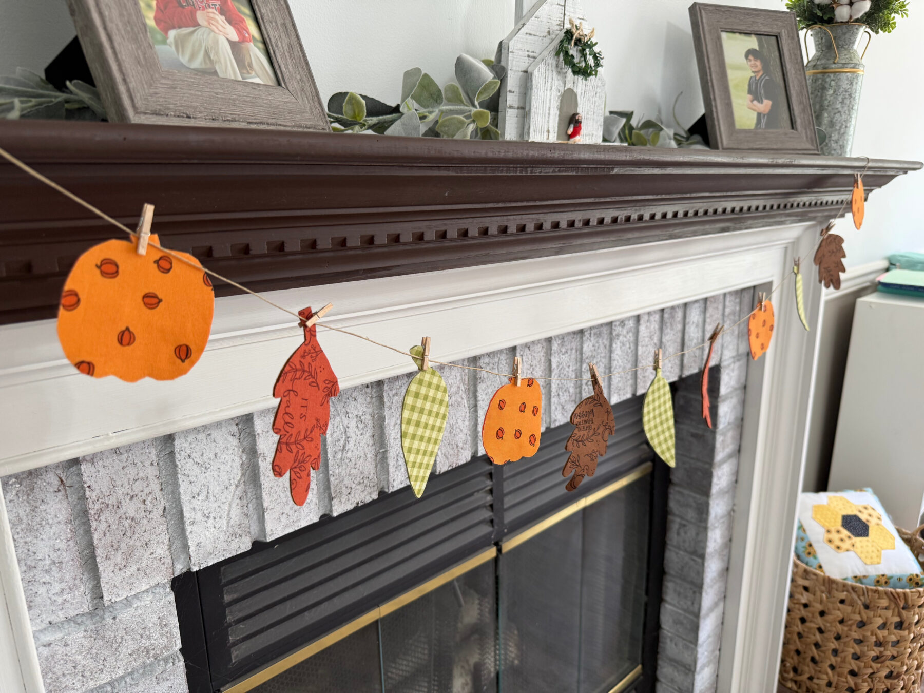 Image contains a decorative harvest banner made of fabric and felt leaves draped across a white fireplace with a dark brown mantel.