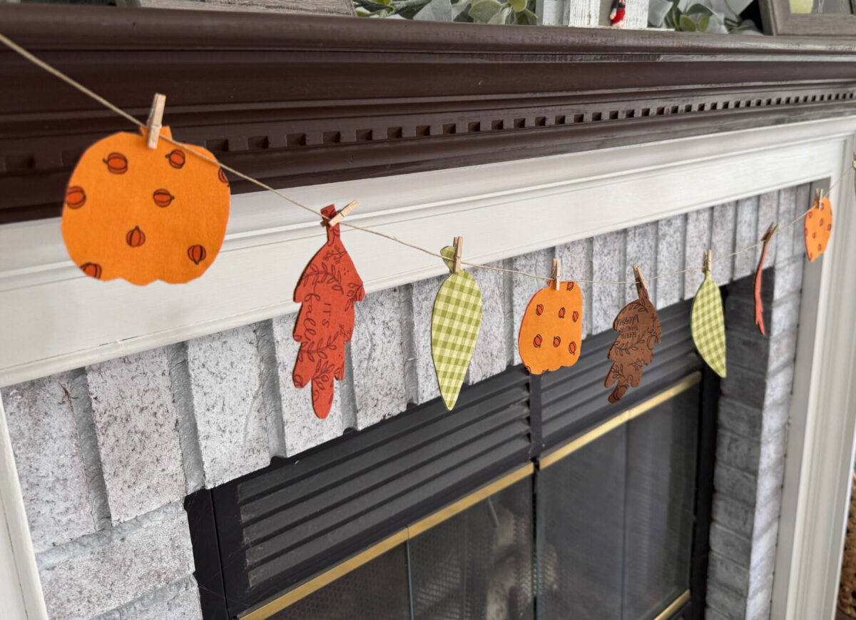 Image contains a fall banner made of fabric leaf and pumpkin shapes clipped to a piece of twine.