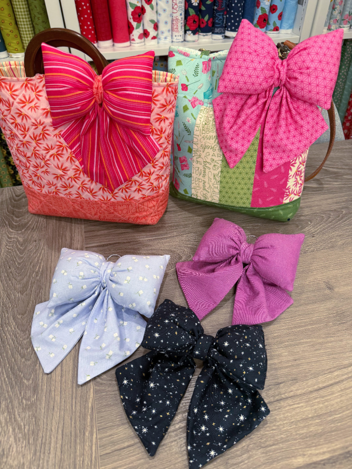 Image contains multi-colored bows made from fabric. Some are clipped to bags while others sit on a wooden desk.