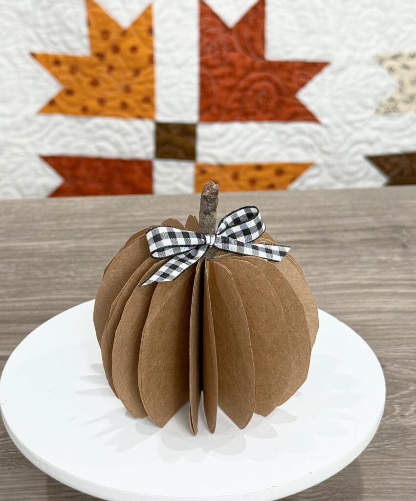 Image contains a paper bag pumpkin sitting on a white round stand on a wooden desk. A fall quilt hangs behind it.
